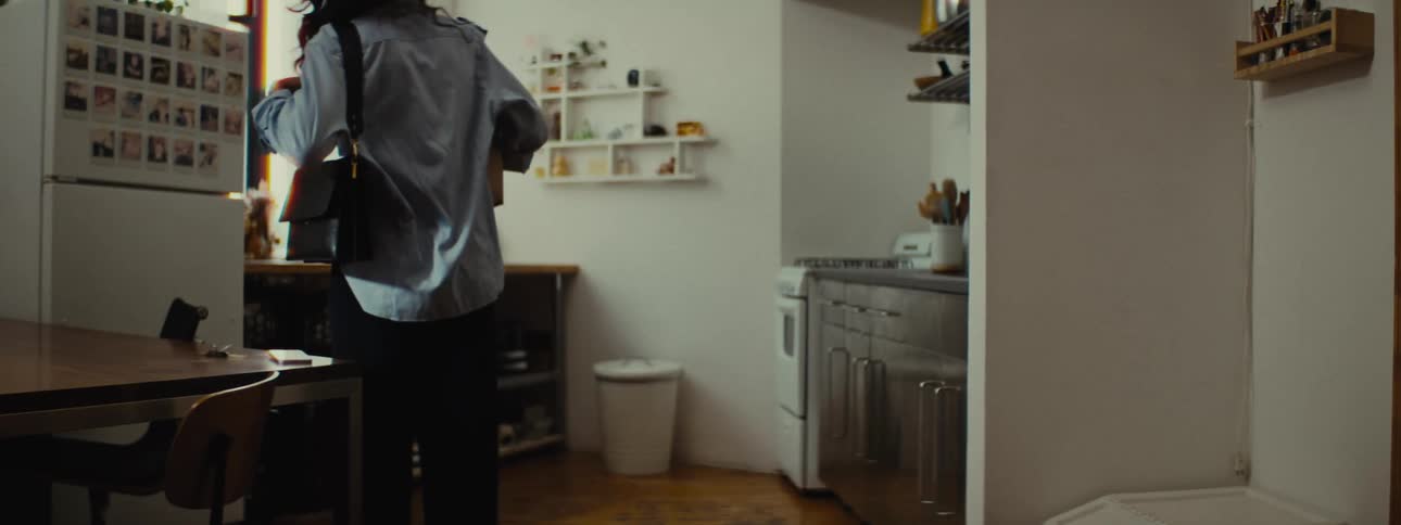 A woman enters her apartment with a grocery bag and mail standing in the kitchen and leaning on a shelf while reading the mail