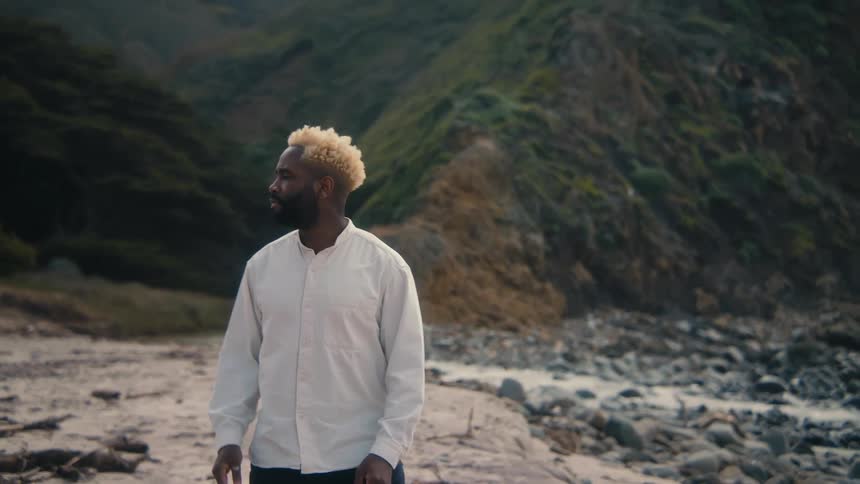 Man in a long white shirt walking on a beach with mountains in the background