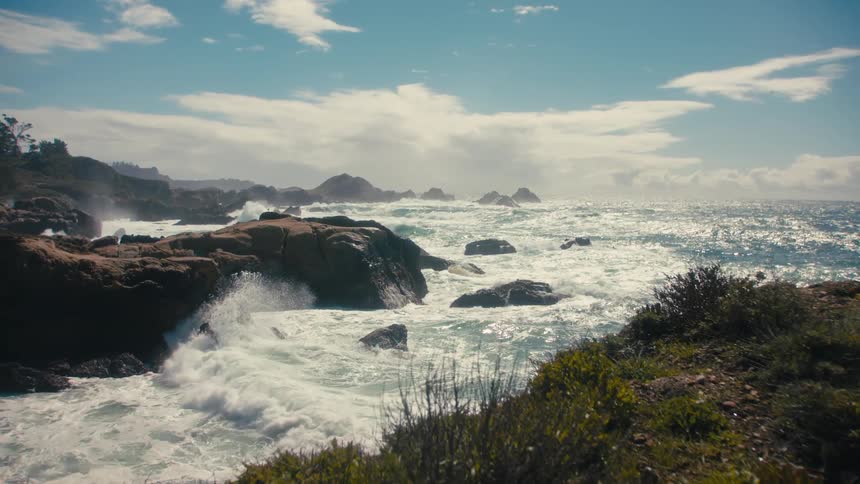 Waves crashing hard into a rocky coastal shoreline cliffs on a bright day under a blue sky