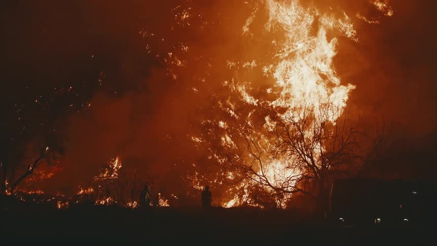 A massive fire rages lighting up the night sky with orange flames and casting the silhouettes of firefighters in the foreground