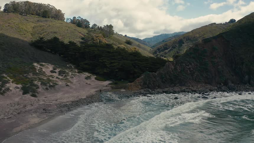 Aerial of ocean waves rolling into a mountainous coastline on a mostly cloudy day