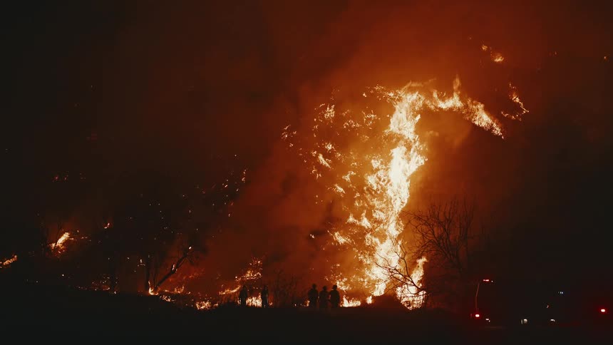 Silhouetted figures stand amidst a raging fire that illuminates the night sky engulfing a large area with flames and smoke