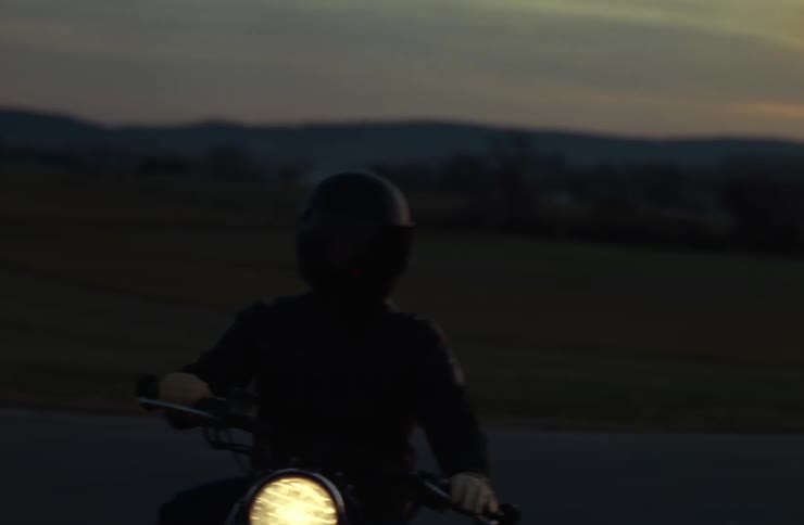 Person dressed in black and wearing a dark helmet rides a motorcycle with the headlight on