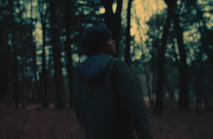 A boy wearing a dark jacket and a beanie is walking through a forest with tall trees during dawn or dusk
