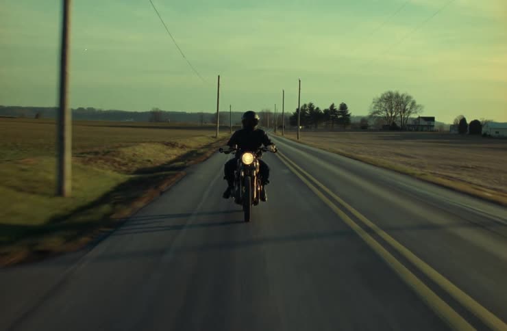A person is riding a motorcycle down a long straight rural road with fields on either side and a fence to the left