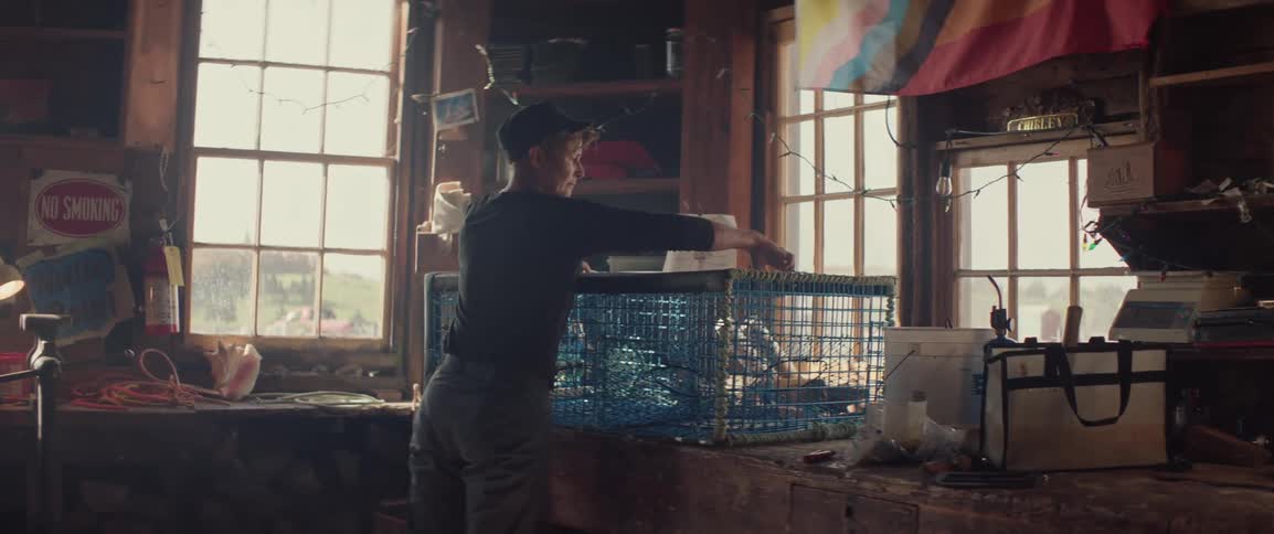 A person stands in a rustic workshop with large windows working on a lobster trap