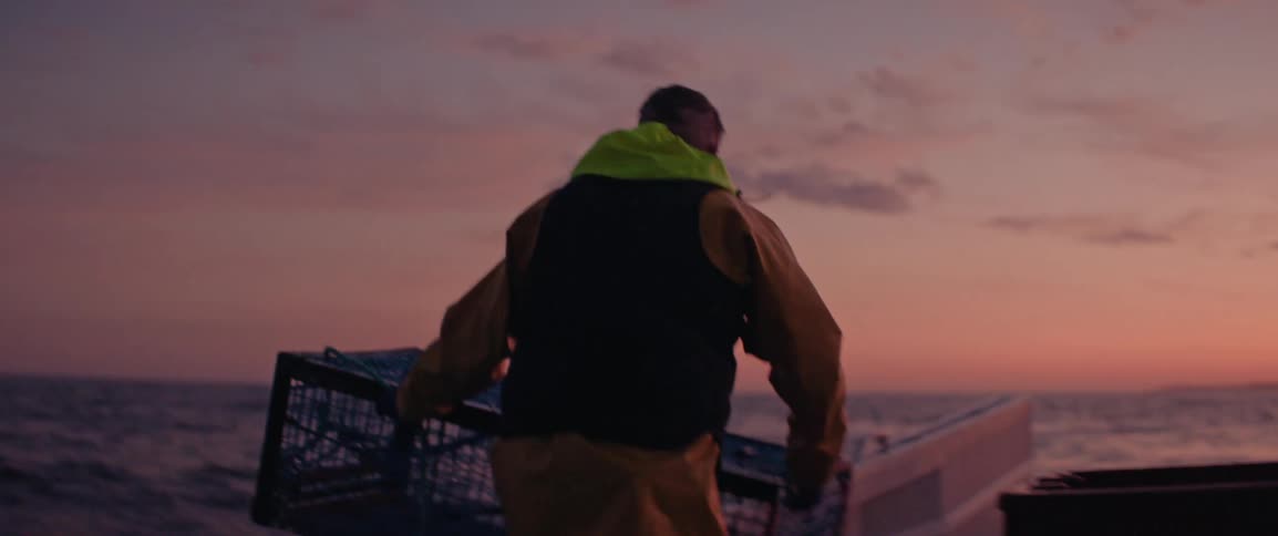 A person in a yellow raincoat and neon green hood places a large rectangular wire lobster cage onto the deck of a boat at sunset