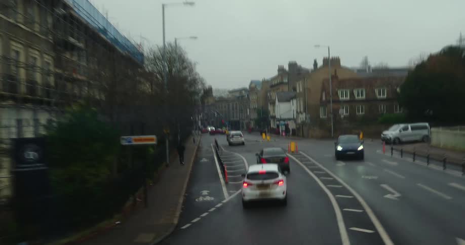 A rainy overcast urban street scene with cars driving along a wet road their red taillights glowing while traffic lights display green signals leafless trees line both sides of the road and a distant hilly neighborhood with houses is visible