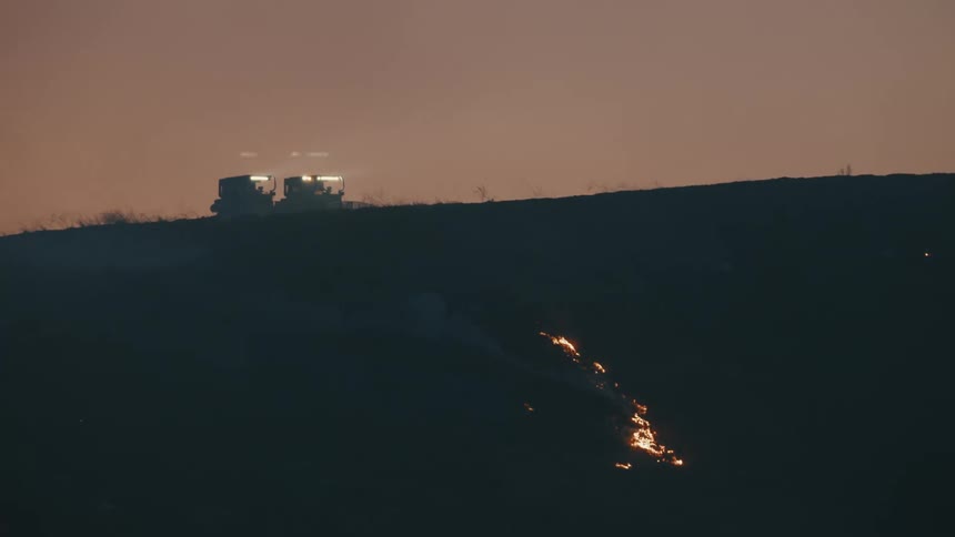 Two vehicles parked on a dark hill beside a bright fiery trail descending a slope likely a wildfire