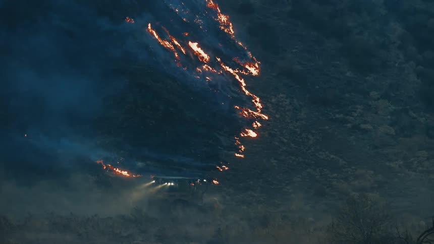 Forest fire consumes a mountainside as a vehicle with headlights on approaches