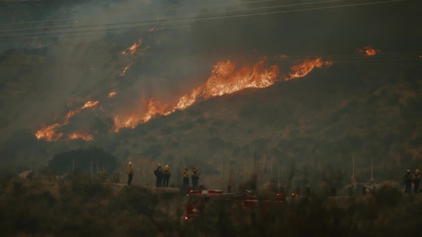 An intense fire burning in the vegetation of a hill with some firefighters at the bottom watching