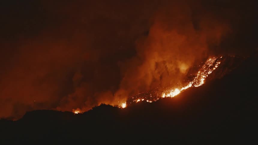 Flames engulf a mountainside in a dark landscape creating a bright orange blaze against the night
