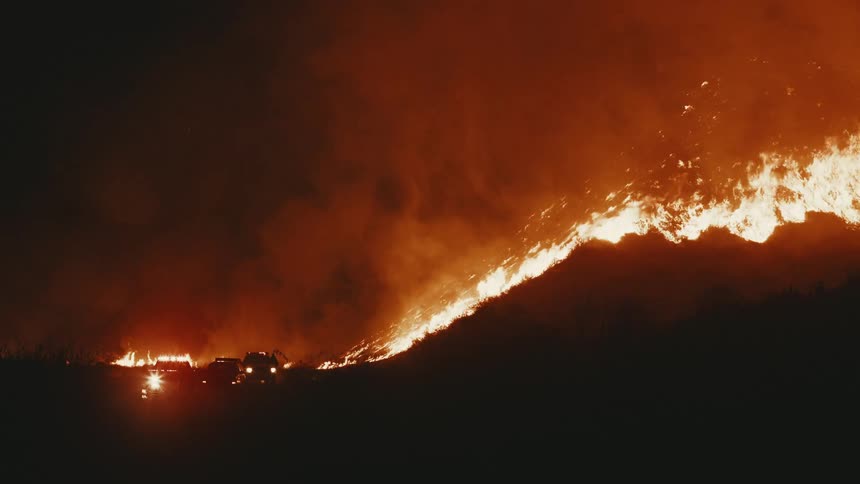 A raging forest fire climbs the hillside at night as silhouetted fire trucks pull up with a firefighter standing near the base illuminated by flashing lights