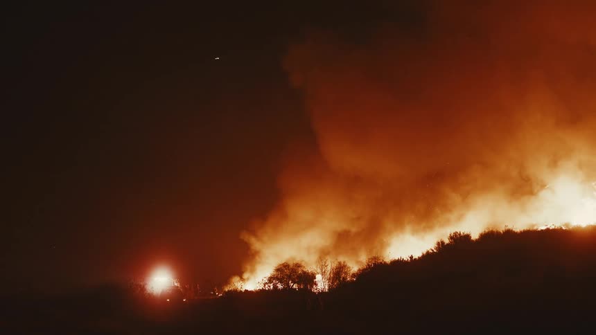 Intense flames and a thick cloud of smoke cover vegetation as a fire rages across a hillside at night
