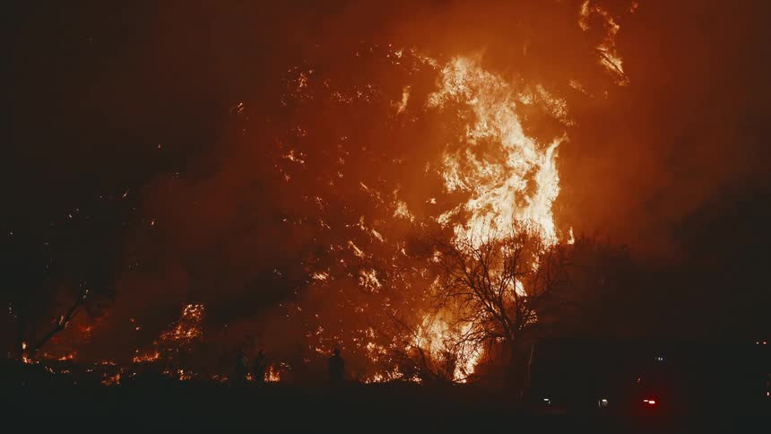 A hillside is engulfed in towering flames shooting skyward as silhouetted firefighters and fire trucks with flashing lights line the city street