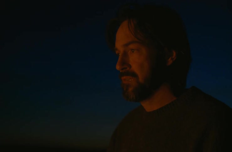 A bearded man kneels in front of a campfire at night