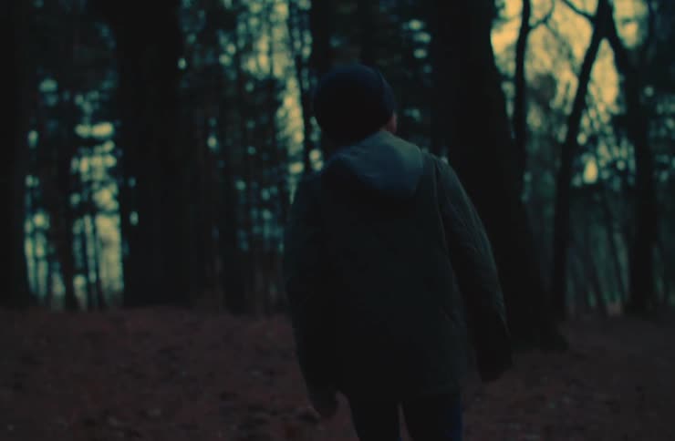 A person walks through a dense forest at dusk wearing a beanie and jacket
