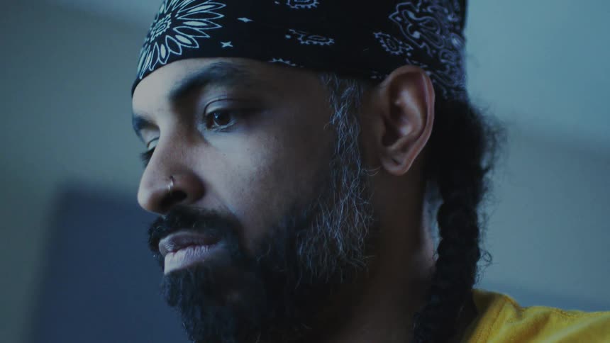 A man with a contemplative expression wearing a black bandana with white patterns his gaze directed downward in a cool toned setting