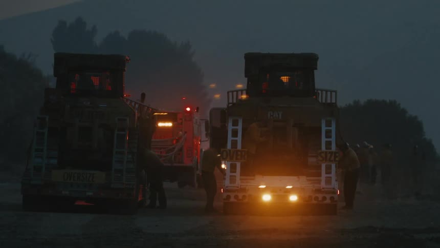 Two large firetrucks with illuminated details stand in a line on a dimly lit hazy landscape with firefighters working between them