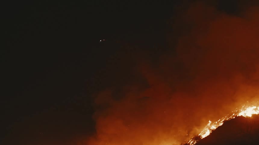 A blazing wildfire engulfs a hillside at night sending thick smoke billowing into the darkness