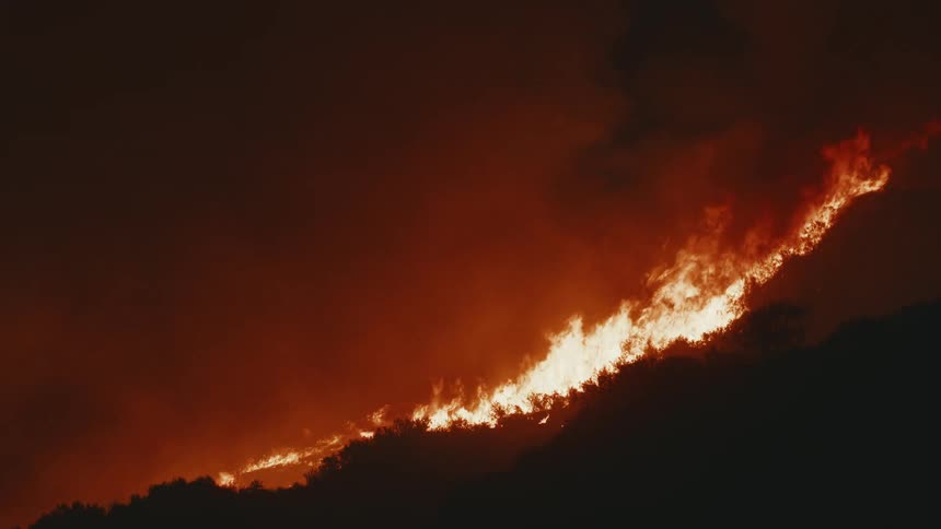 A dark silhouetted hillside is ablaze with a long bright orange fire line against a smoky reddish sky