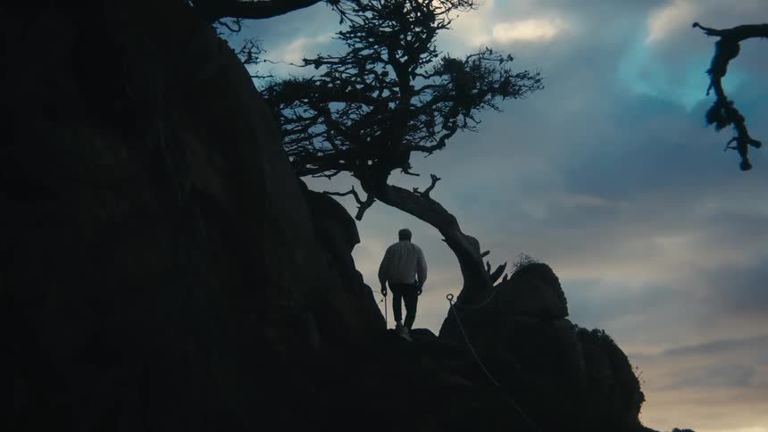 Man walking up a rock hillside staircase path in front of a dimly lit blue and cloudy sky background