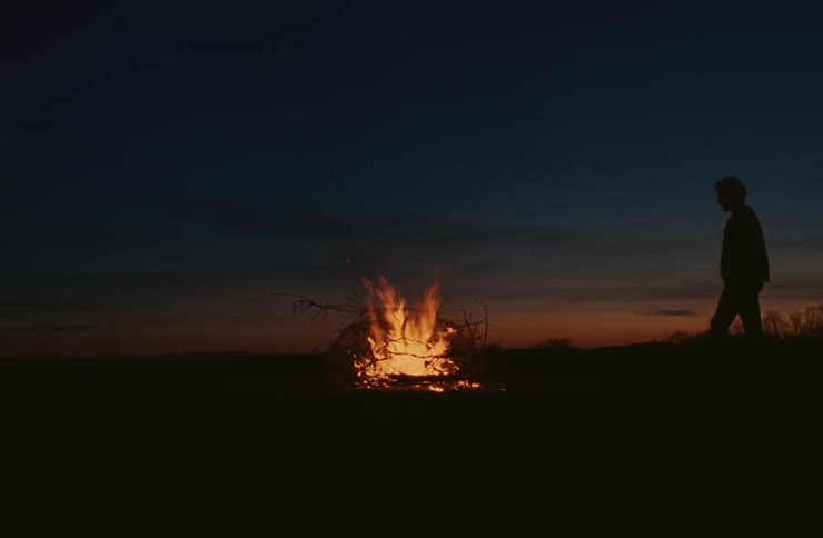 A solitary figure stands silhouetted against a dark landscape watching a small fire burning in the foreground