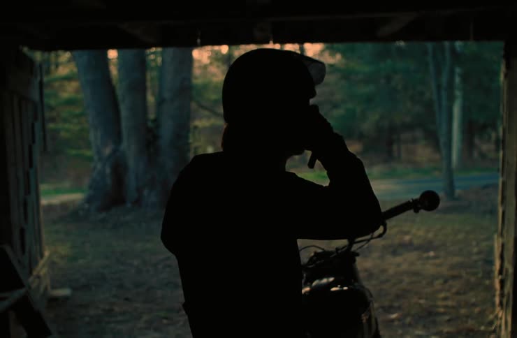 The silhouette of a person putting on a helmet and then starting a motorcycle in an environment where the front looks out onto a forest