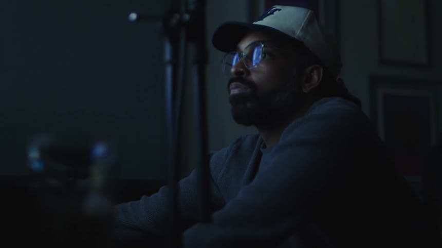 A person wearing a cap and a hoodie is sitting in a dimly lit room with a microphone stand in the foreground