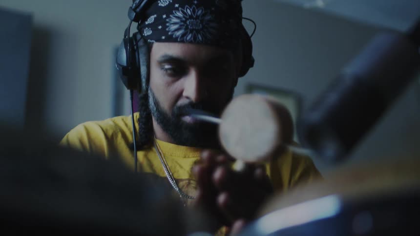 A bearded man wearing a patterned bandana and headphones deeply focused as he plays a percussion instrument in a dimly lit recording studio a microphone is visible in the foreground capturing the sound while the background remains softly blurred