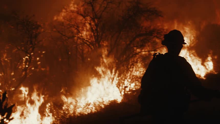 A silhouetted firefighter stands amid a raging fire with flames engulfing the surrounding area