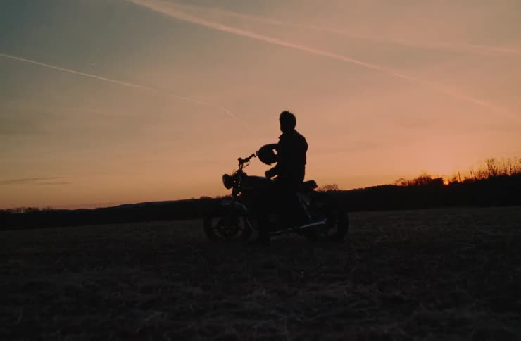 A silhouette of a person sits on a motorcycle gazing at the horizon as the sun sets casting a warm glow over the sky