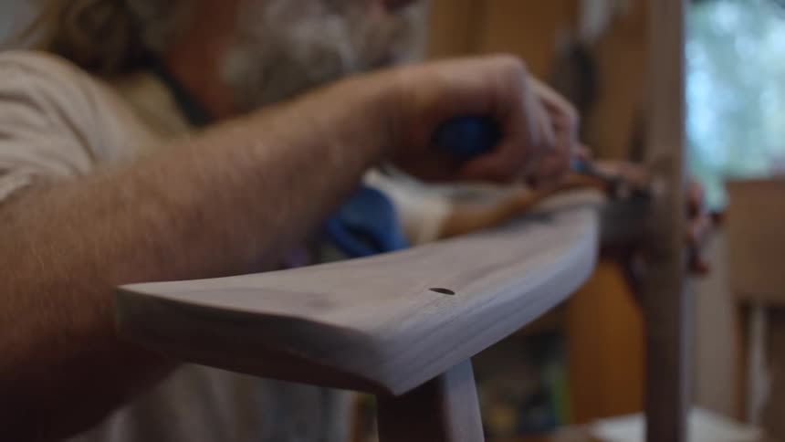 The face of a grayhaired bearded man who is working with a tool on a wooden object
