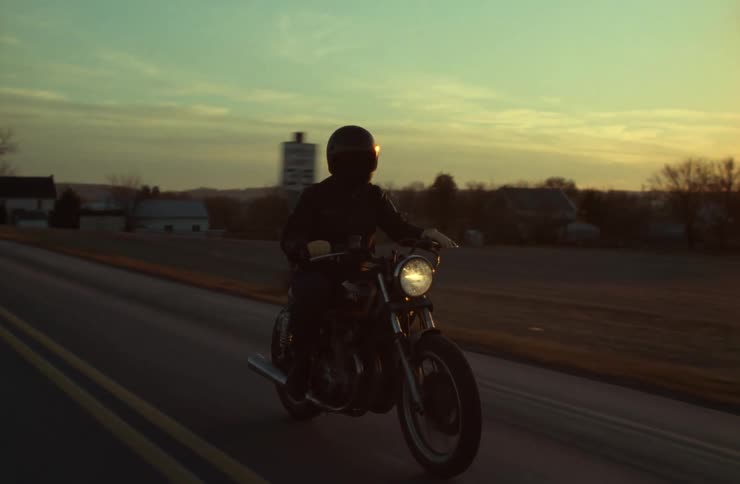 An adult rides a motorcycle on a rural highway at sunset