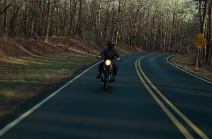 A motorcyclist rides down a winding doubleyellowlined road through a forest with bare trees