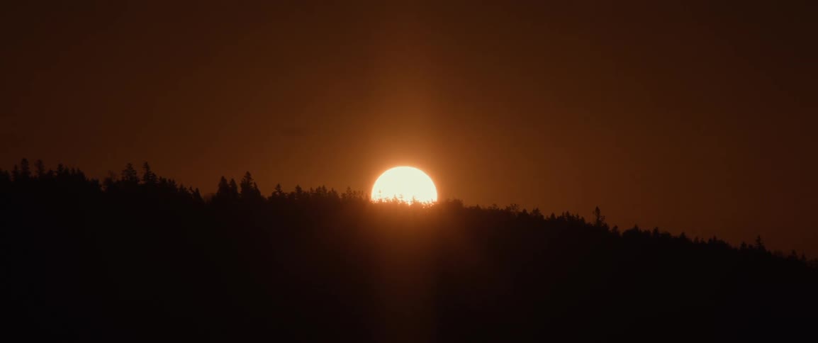 Zoom out of the sun setting behind a forested ridge