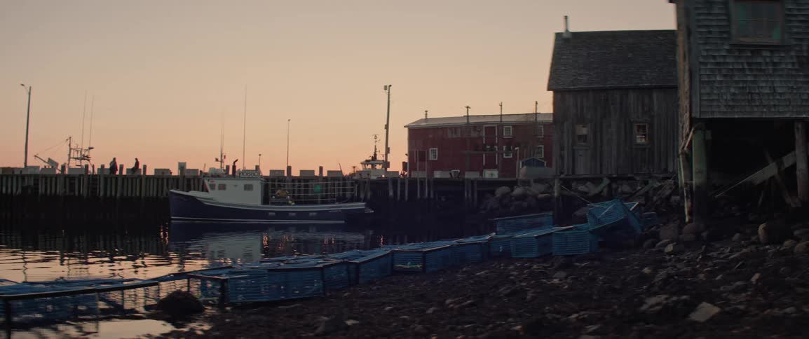 A fishing boat sits moored in the calm waters of a coastal town as crab traps lie along the shore under a soft morning sky