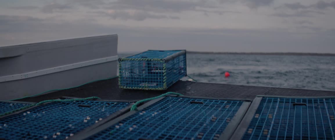 A lobster trap is seen tipping over on the deck of a boat with the open ocean and a cloudy sky in the background