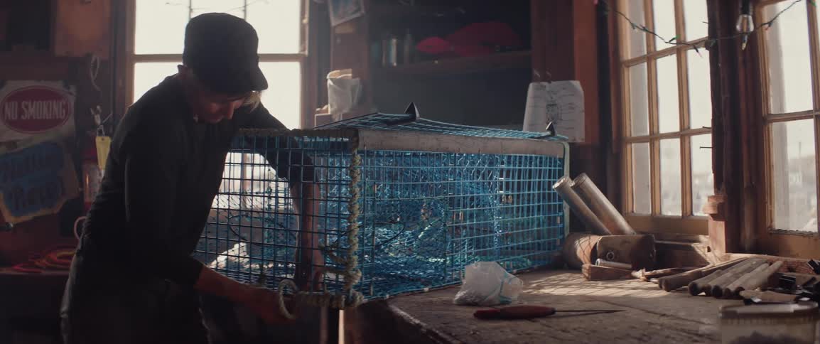 Person adjusting a blue fishing trap inside a wooden workshop lit by window light