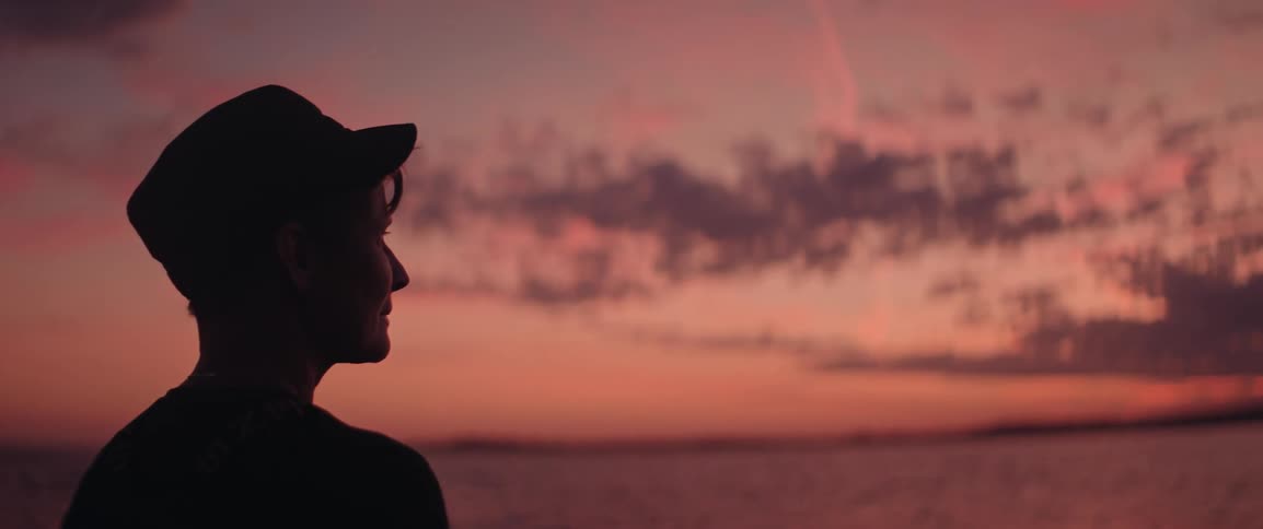 Woman wearing a dark cap looks out to sea under an orange and pink sky at dusk
