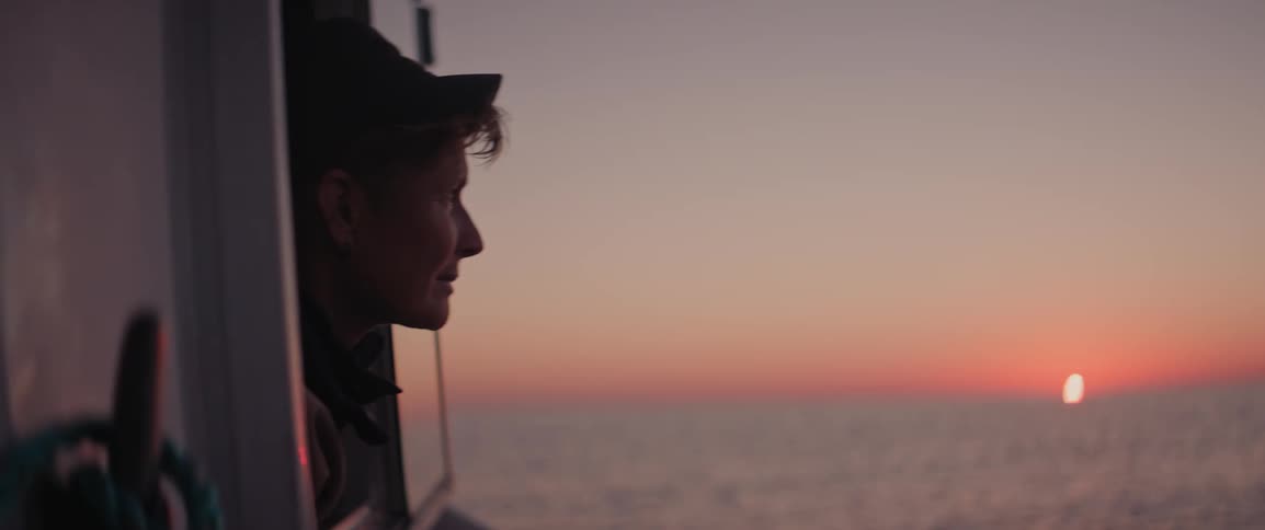 Person looking out the window of a boat moving across the sea with the sunrise on the horizon