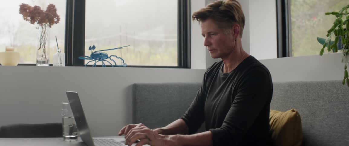 A woman sits indoors at a table and types on her laptop