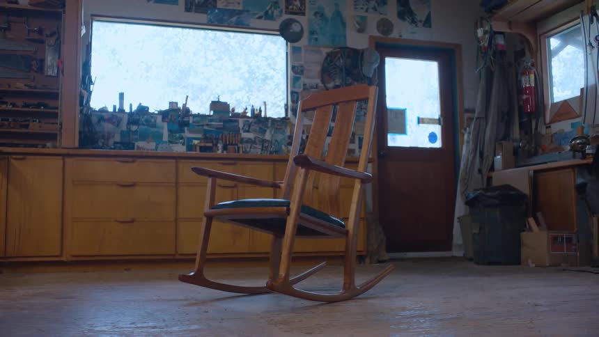 A wooden rocking chair with a blue padded seat sits in a cluttered room with a brightly lit window and various items on the walls
