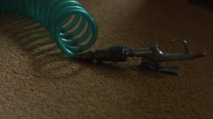 A coiled green air hose lies on a brown carpet next to a silver metal spray gun