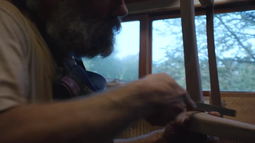 A person is working on a wooden chair in a cluttered workshop with various tools materials and equipment visible