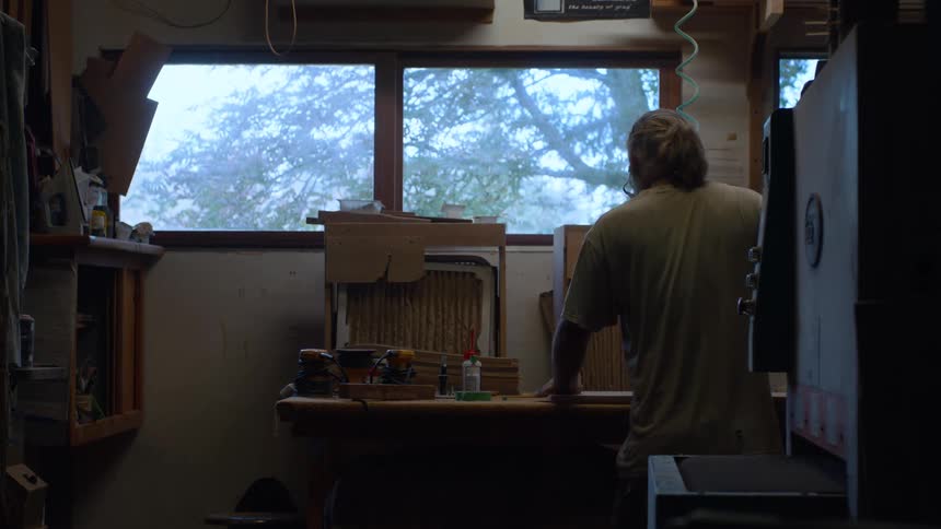 A person works at a woodworking workshop next to a window in a dimly lit room