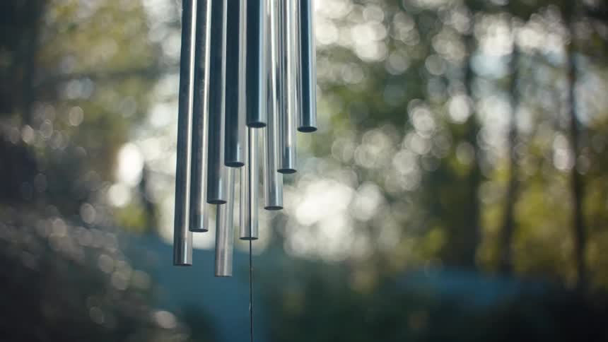 Metal tubes of a wind chime hang against a blurred background of trees illuminated by sunlight