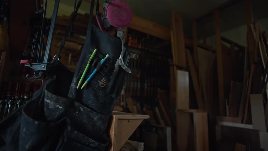 A cluttered woodworking shop with various tools and materials including a black apron hanging with pencils and a measuring tape in its pockets and wooden boards stacked in the background