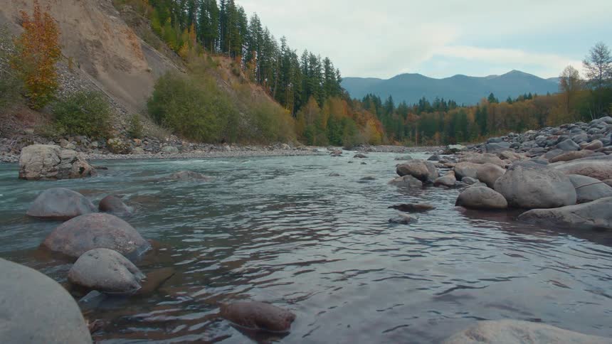 At river level the mountain river flows by slopes of autumn trees with distant peaks rising on the horizon