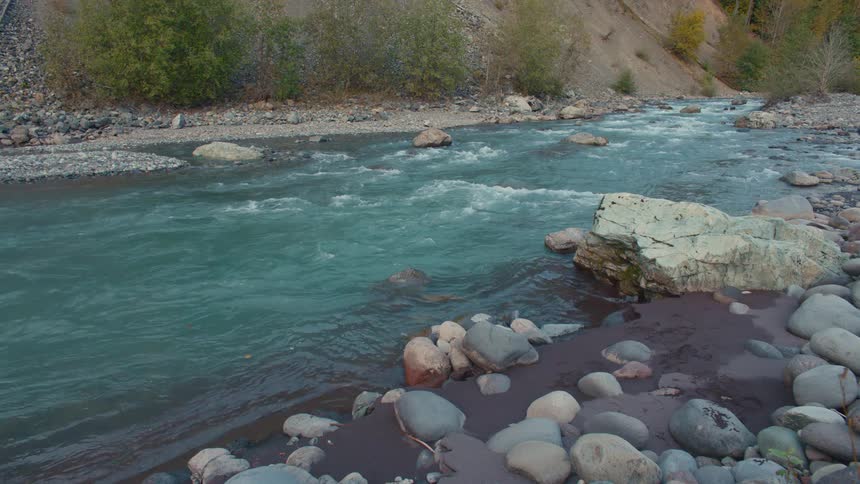 River with blue waters runs between stones and rocky banks surrounded by vegetation and slopes